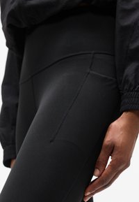 Εγγύτατη λήψη του χεριού ενός ατόμου που ακουμπά στο πλευρό του, φορώντας μαύρο κολάν με ψηλή μέση και μαύρο μακρυμάνικο τοπ με ελαστικά μανσέτα.