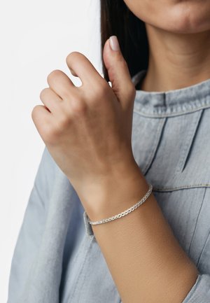 Bracciale d'argento con forme ovali collegate, caratterizzato da una finitura liscia. Indossato su un polso, su uno sfondo di tessuto di colore azzurro chiaro.