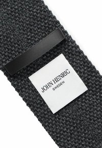 Mörk texturerad tyg med ett vävt mönster, prydd med en vit etikett med svart text "JOHN HENRIC SWEDEN" och en svart satinrem.