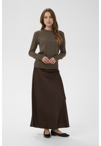 Brauner Strickpullover mit gerippten Bündchen, kombiniert mit einem langen, glatten, dunkelbraunen Satinsrock. Das Modell trägt schwarze flache Schuhe und steht vor einem neutralen Hintergrund.