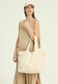 Beżowa torba płócienna z dwoma uchwytami u góry, przednimi kieszeniami, białymi akcentami i nadrukowanym tekstem "MELBOURNE TENNIS CLUB" w okrągłym wzorze.