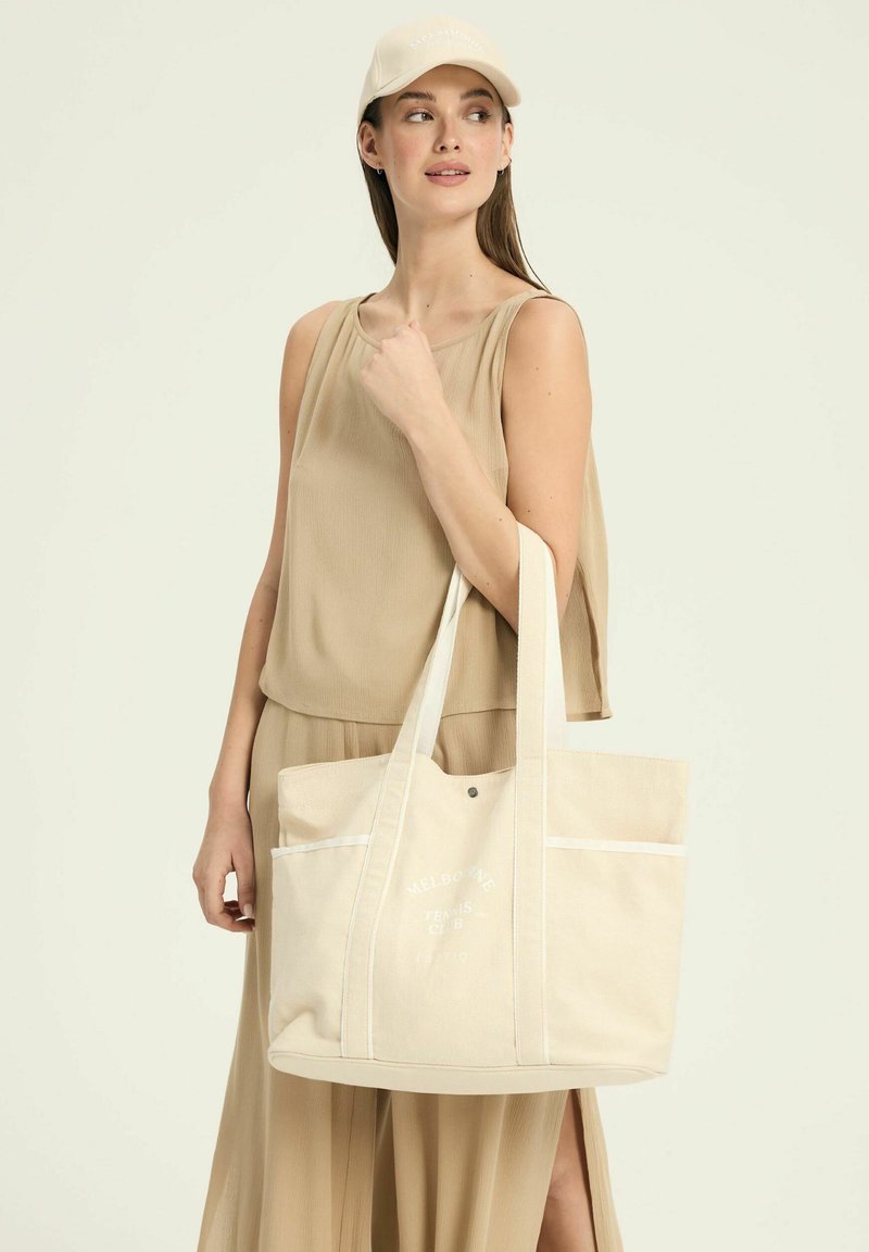 Beżowa torba płócienna z dwoma uchwytami u góry, przednimi kieszeniami, białymi akcentami i nadrukowanym tekstem "MELBOURNE TENNIS CLUB" w okrągłym wzorze.