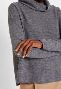 Tröja med kappa-hals i en texturerad grå och svart stickning, med långa ärmar och en avslappnad passform. Nederkanten är rak med en något lös skärning.