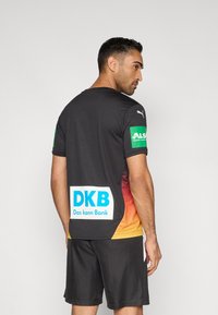 Black athletic shirt with gradient orange-yellow accents. Features logos on sleeves and back, short sleeves, and lightweight fabric.