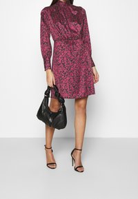 Robe à motif fuchsia à manches longues et col haut, assortie à un sac à main en cuir noir et des talons noirs à brides.