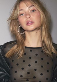 Crna mrežasta majica s točkicama i okruglim izrezom, nosi se ispod crne kožne jakne, s dugom plavom kosom i istaknutim srebrnim naušnicama u obliku obruča.