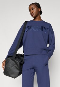 Tmavě modrá mikina DKNY s vyraženým logem, volným střihem a žebrovaným lemem; doplněná o sladěné tepláky. Modelka drží černou sportovní tašku.