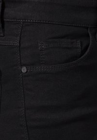Jeans de mezclilla negra con una textura suave. Incluye un pequeño bolsillo frontal y costuras negras. El material es elástico y ajustado.