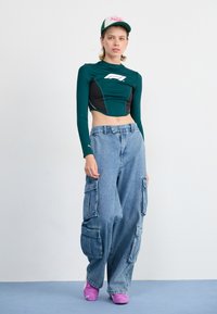 Groene lang-slavend cropped top met zwarte accenten, blauwe baggy cargo jeans en roze instap schoenen. Witte achtergrond, minimalistische setting.