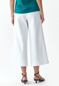 Bílé široké kalhoty s hladkou texturou, doplněné černými sandály na tenkých podpatcích s malým podpatkem, stojící na neutrálním pozadí.