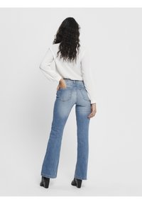 Nainen, jolla on pitkät kiharat hiukset, pukeutuneena valkoiseen paitaan, vaaleansiniset bootcut-farkut ja mustat korokepohjaiset saappaat, seisoo yksi käsi takataskussa.