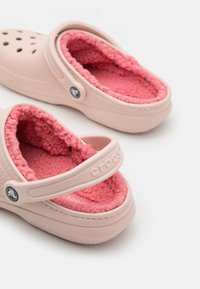 Sabots rose clair avec une doublure douce et duveteuse de couleur rose. Caractéristiques d'une sangle, d'un dessus perforé pour la respirabilité et d'une semelle texturée pour une meilleure adhérence.