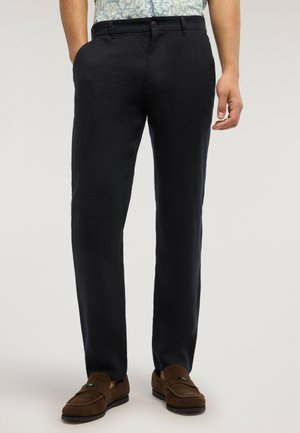 Mand iført sorte skræddersyede bukser og brune ruskindsløvesko, stående med den ene hånd afslappet ned langs siden.