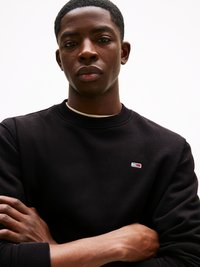 Sweatshirt negru cu manșete și guler ribate, având un mic logo pe pieptul stâng. Model cu brațele încrucișate, purtând un tricou de culoare deschisă.