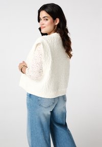Witte textuurvest met hoge nek, mouwloos ontwerp en decoratief patroon, gecombineerd met lichtblauwe wijde spijkerbroek en een kanten top.