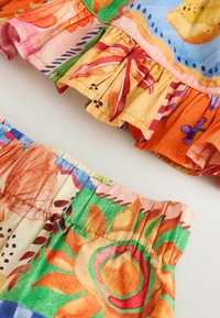 Tissu coloré vif avec taille élastique froncée et ourlet volanté, présentant des motifs floraux et abstraits dans des nuances d'orange, de vert, de bleu et de rose.