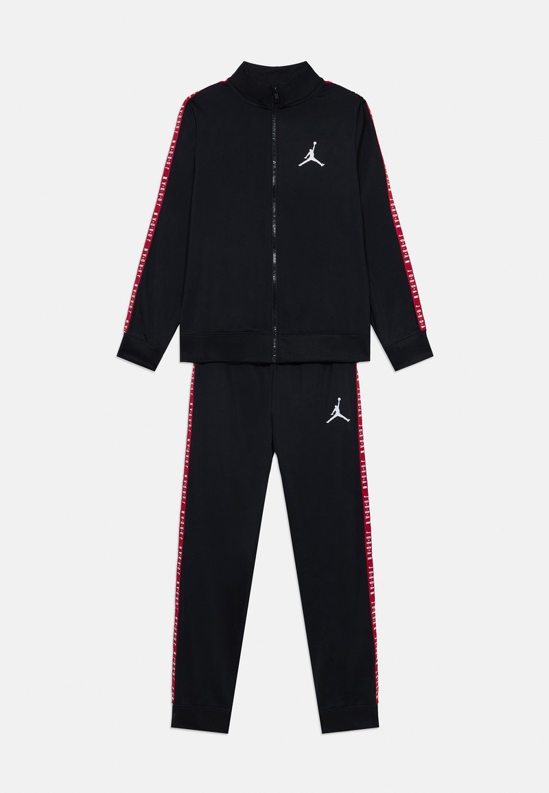 Survêtement noir en tissu lisse, comprenant une veste zippée avec des rayures latérales rouges et un logo blanc, associée à un pantalon assorti.