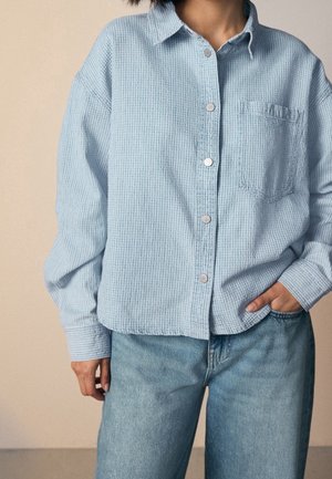 Person wearing a light blue textured button-up shirt with a chest pocket and light blue jeans, one hand in pocket, neutral background.