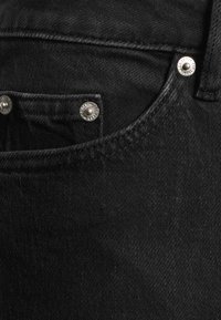 Vaqueros de mezclilla negra con un bolsillo frontal, remaches de tono plateado y detalles de costura sutiles. La tela tiene un aspecto suave y texturizado.