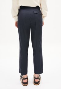 Persona vestida con pantalones rectos azul marino y sandalias negras con forro de piel sintética beige, de pie contra un fondo blanco liso.