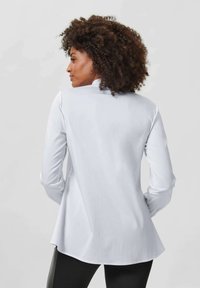 Wit, lang mouwshirt met een gebogen zoom en een gladde textuur. Voorzien van een hoge kraag en aansluitende mouwen, gestyled met zwarte leggings.