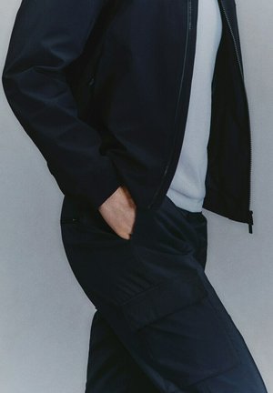 Persona con chaqueta negra con cremallera y pantalones cargo negros, con la mano en el bolsillo, camisa de color claro visible debajo de la chaqueta.