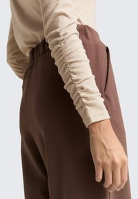 Bruine stoffen broek met steekzakken, gecombineerd met een licht beige top met lange mouwen en gerimpelde mouwdetails.