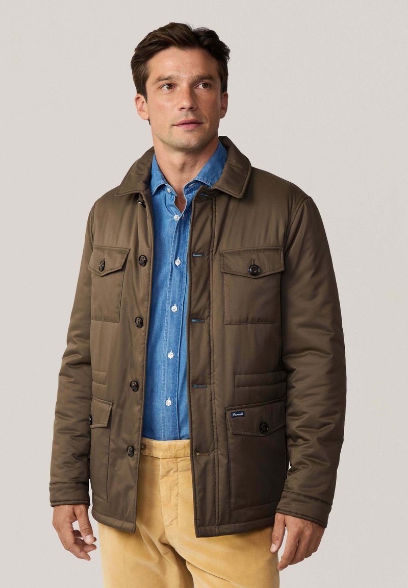 Chaqueta aislante marrón con cuello, múltiples bolsillos frontales y cierres de botones. Llevada sobre una camisa de mezclilla y pantalones beige.