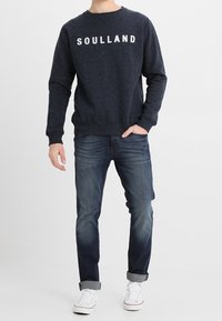 Felpa navy con il testo bianco "SOULLAND", scollatura a girocollo, tessuto testurizzato. Abbinata a jeans blu scuro e sneakers bianche.