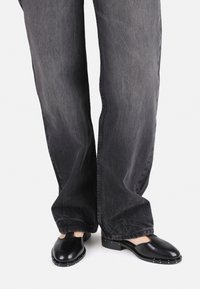 Schwarze Lederclogs mit Ausschnittmuster und metallischen Nieten. Kombiniert mit dunkelgrauen, lässig geschnittenen Jeans. Fokus auf die Schuhe und die unteren Details der Hose.