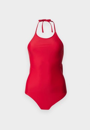 Femme debout pieds nus portant un maillot de bain une pièce rouge sans manches avec un col montant sur un fond clair uni.