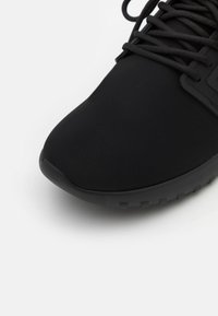 Chaussure de sport noire avec une tige en tissu lisse et texturé, un bout arrondi, et une semelle en caoutchouc présentant un motif de bande de roulement.