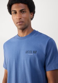 Blauwe katoenen T-shirt met een ronde hals en korte mouwen. Op de voorkant staat de tekst "ANTICO BAR by MoPD" in contrasterende kleuren.
