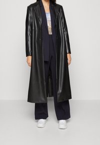 Persoon in een lange zwart leren jas over een marineblauw pinstripe pak met een graphic top en witte sneakers, staande tegen een effen lichte achtergrond.