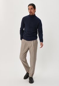 Pulover cu guler înalt bleu închis, pantaloni bej ajustați cu o textură subtilă și pantofi negri cu șireturi. Modelul stă într-o poziție relaxată.