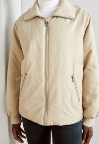 Beige bomberjakke med blød krave, frontlynlås og to sidelommer med lynlås. Glat tekstur og let puffet design.