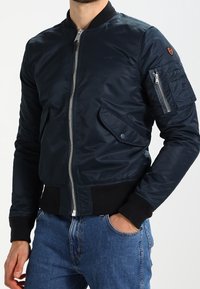 Homme portant une veste bomber bleu marine avec fermeture éclair à l'avant, poches à rabat et poche utilitaire zippée sur la manche gauche, associé à un jean bleu.