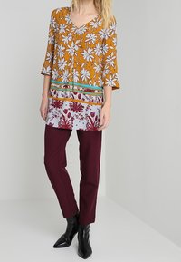 Tunique imprimée en moutarde avec des motifs floraux, associée à un pantalon bordeaux. Présente un décolleté en V et des manches trois-quarts. Bottines noires.