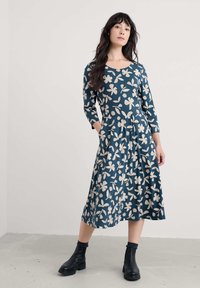 Sukienka o długości do kolan w ciemnym tealowym kolorze z kwiatowym wzorem, z długimi rękawami i dopasowanym gorsetem. Posiada kieszenie i rozkloszowany dół. Na nogach czarne botki za kostkę.