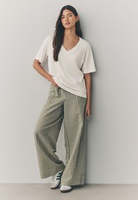 Maglietta a V color crema abbinata a pantaloni a gamba larga a quadretti verdi e bianchi. Il modello indossa sneaker bianche con strisce verdi. Tessuto liscio.