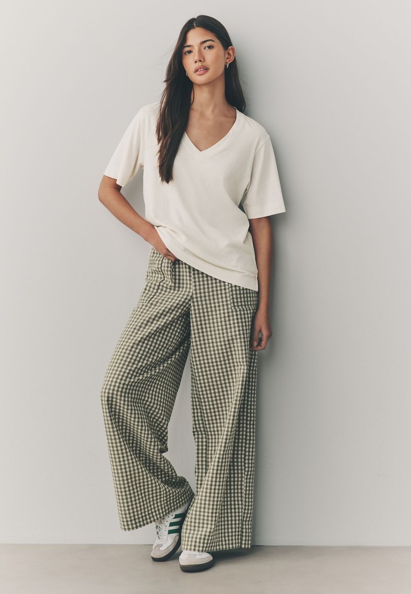 Maglietta a V color crema abbinata a pantaloni a gamba larga a quadretti verdi e bianchi. Il modello indossa sneaker bianche con strisce verdi. Tessuto liscio.