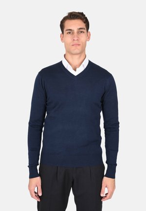 Maglione blu navy a collo a V realizzato in un tessuto a maglia liscia, con maniche lunghe e vita aderente. Indossato sopra una camicia bianca con colletto.