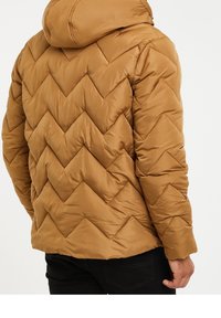 Bruine pufferjack met een chevron doorgestikt patroon, warme stof en een vastgemaakte capuchon, gecombineerd met een zwarte broek.