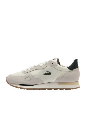 Zapatillas Lacoste con parte superior de lona beige claro, detalles en ante, lengüeta del talón verde y suela de goma. Logo de cocodrilo bordado en el lateral.