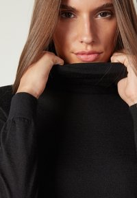 Schwarzer Rollkragenpullover mit einem strukturierten Strickdesign. Nahaufnahme, die den hohen Kragen zeigt und Hände, die ihn sanft nach oben ziehen. Glattes Material.