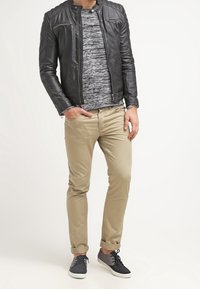 Homme portant une veste en cuir noir, une chemise à motifs gris, un pantalon beige retroussé, et des baskets noir et gris, se tenant avec une main dans la poche.