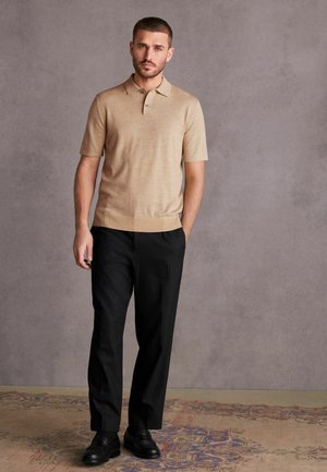Beige kortærmet polo shirt med krave, parret med sorte skræddersyede bukser og sorte sko, stående på et mønstret tæppe mod en grå baggrund.