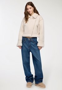 Giacca in shearling beige chiaro tagliata, con maniche larghe, tasche con cerniera e colletto, abbinata a jeans in denim blu larghi e calzature beige.