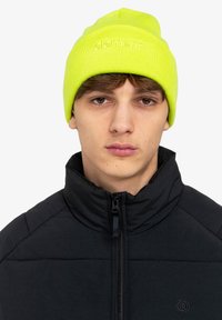 Berretto di maglia gialla con texture a coste e logo "element" ricamato sul fronte, indossato con una giacca nera con cerniera.
