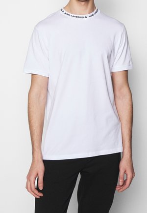 Mann trägt ein schlichtes weißes T-Shirt mit kurzen Ärmeln und dem Schriftzug "KARL LAGERFELD" am Rundhalsausschnitt, kombiniert mit schwarzen Hosen, vor weißem Hintergrund.